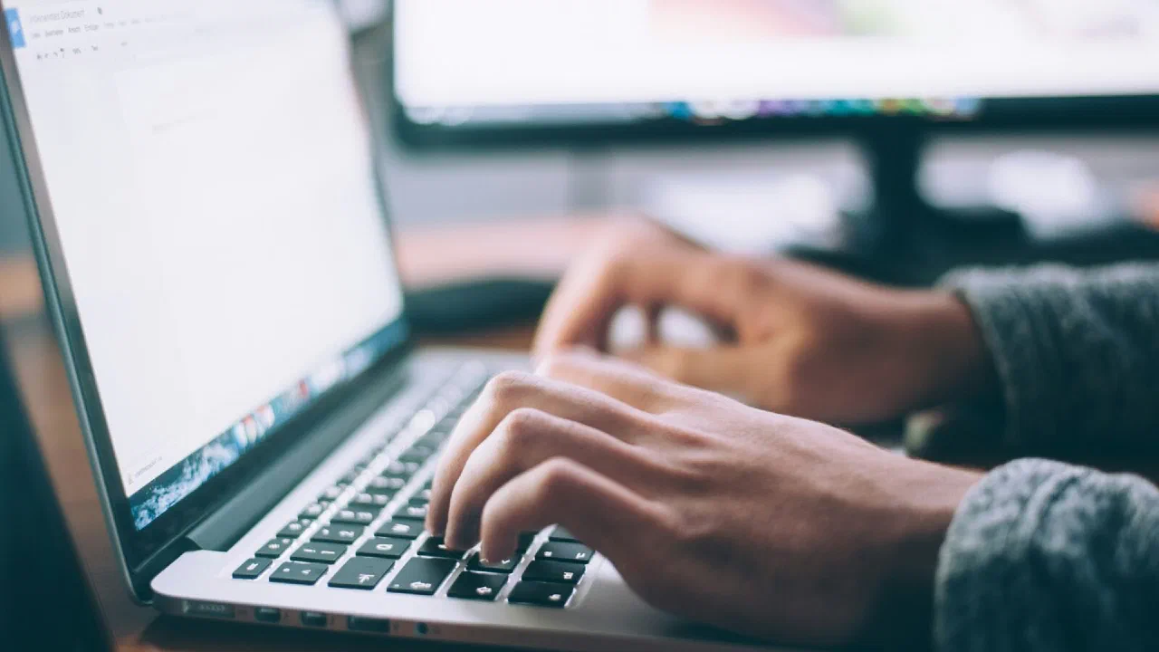 hands typing on a laptop beside a notebook and pen